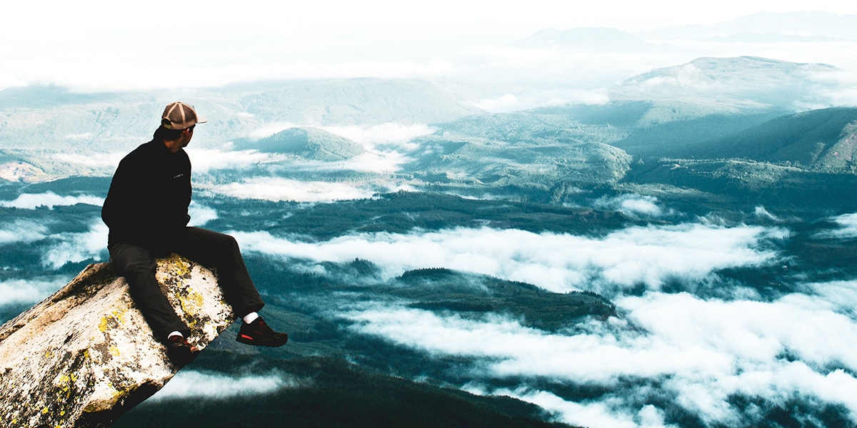 man sitting on a rock ledge surrounded by mountains and deep valleys.