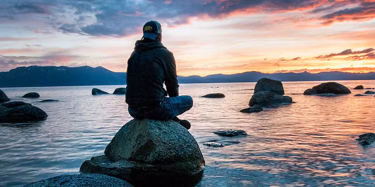Young man sitting on a rock in water and watching a sunset, seeking serenity