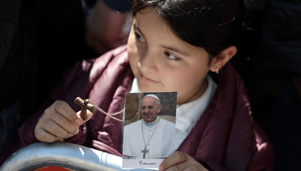 Una niña sostiene una cruz y una foto del Papa Francisco mientras espera la llegada del féretro del Papa Francisco antes de su la Misa exequial en la Plaza de San Pedro en el Vaticano el 26 de abril de 2025. (Foto OSV News/Mohammed Salem, Reuters)